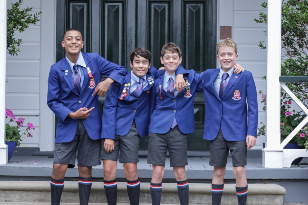 Four boys wearing blue Wellelsey College blazers, ties, and shorts stand smiling together on a porch, arms around each other in front of a green door and white building