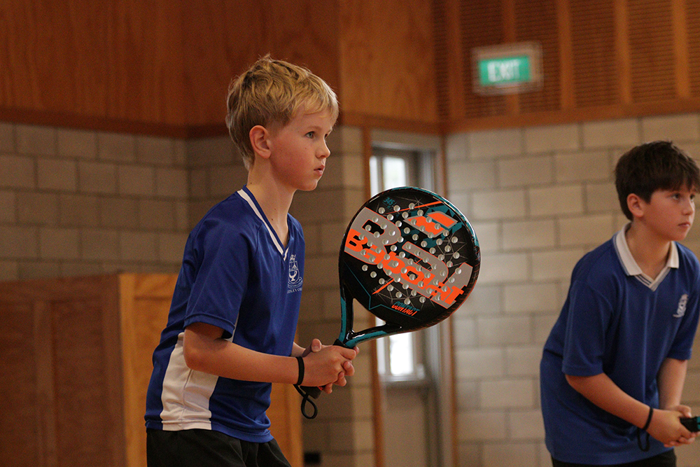 Two boys in blue shirts play paddle tennis indoors. The boy in the foreground holds a paddle, focused and ready, while the second boy stands behind him, also prepared for the game. The setting is a gym with brick and wood walls