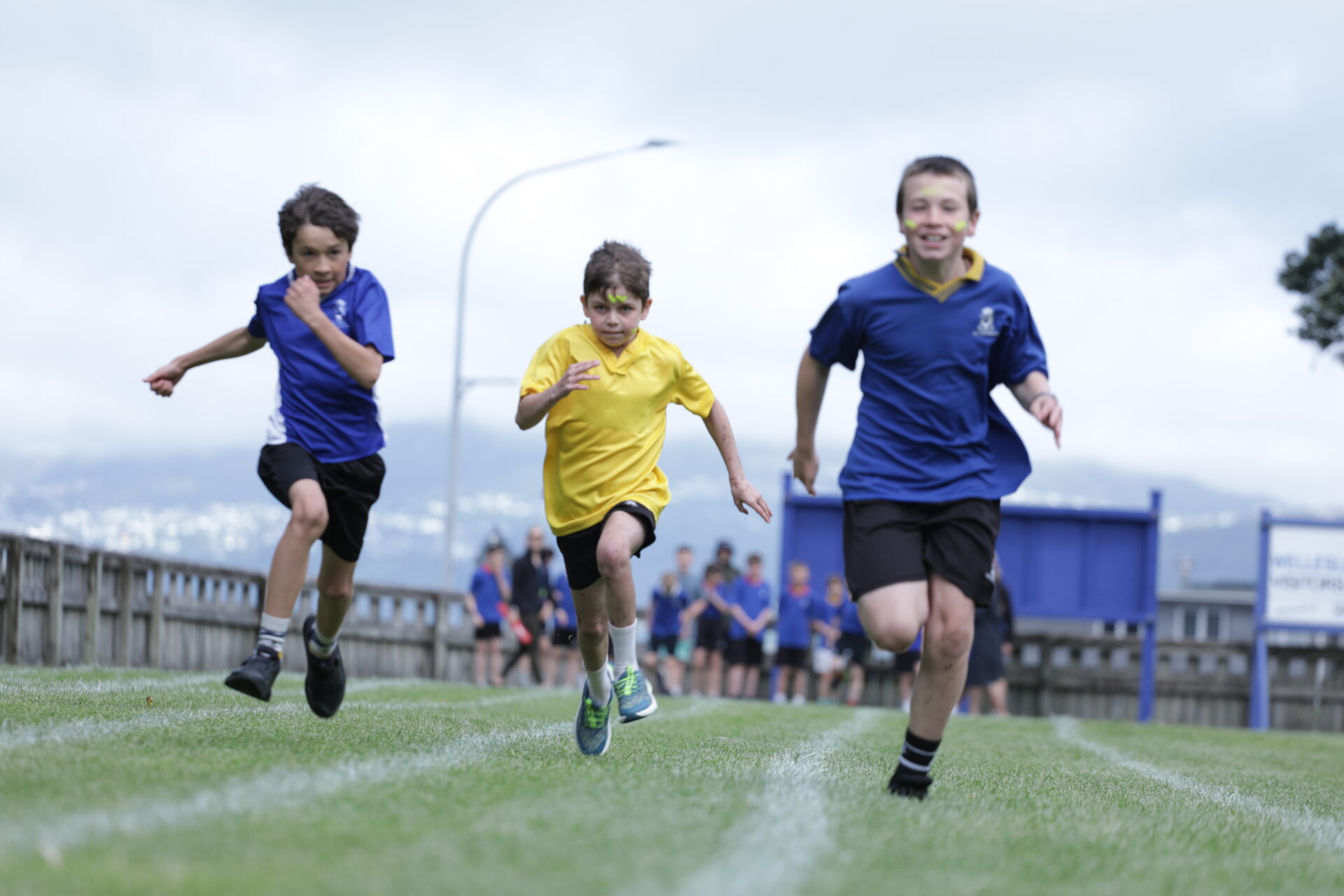 Three boys in Wellesley College sports uniforms race on a grassy track, with one in yellow shirt leading and two in blue shirts following. A group of children and a fence are visible in the background under a cloudy sky
