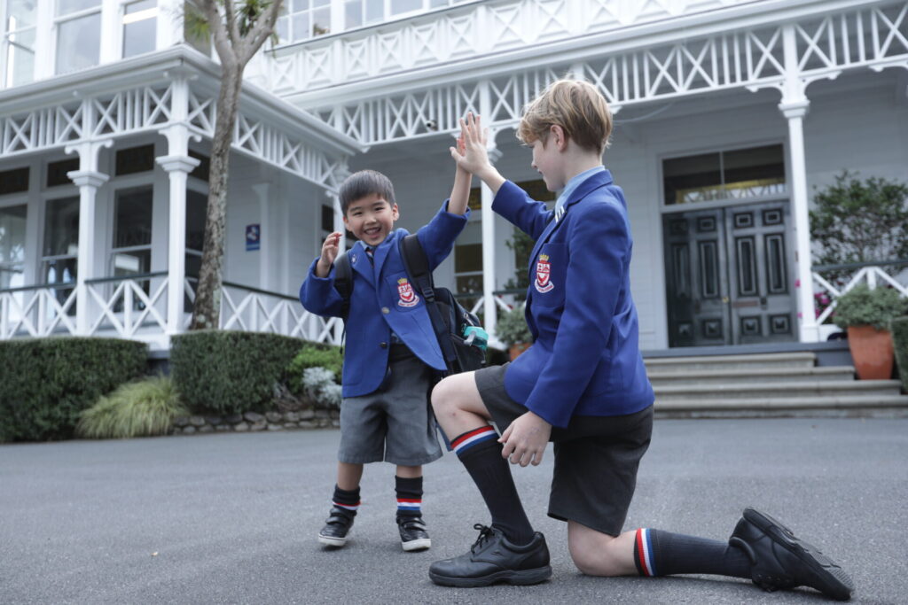 Two schoolboys in blue Wellelsey College blazers and grey shorts give each other a high five outside a large, white building with a porch and steps. One boy is standing and the other is kneeling, both smiling happily