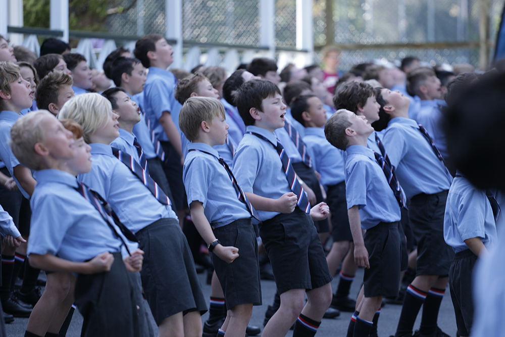 A group of Wellesley College boys performing the haka outdoors, standing in formation with intense expressions and strong, synchronised movements, wearing school uniforms