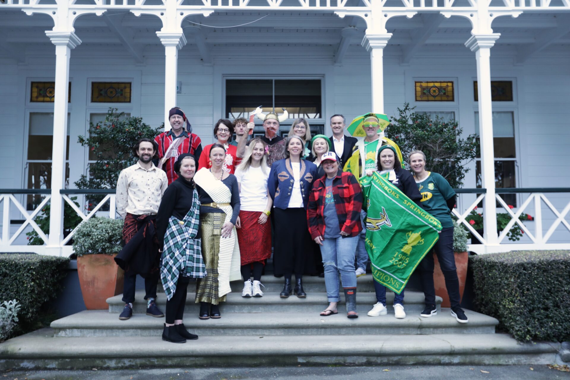 Wellesley College staff in colorful, themed outfits stand and smile on the steps of a white building with decorative trim and large potted plants, suggesting a festive or costume event