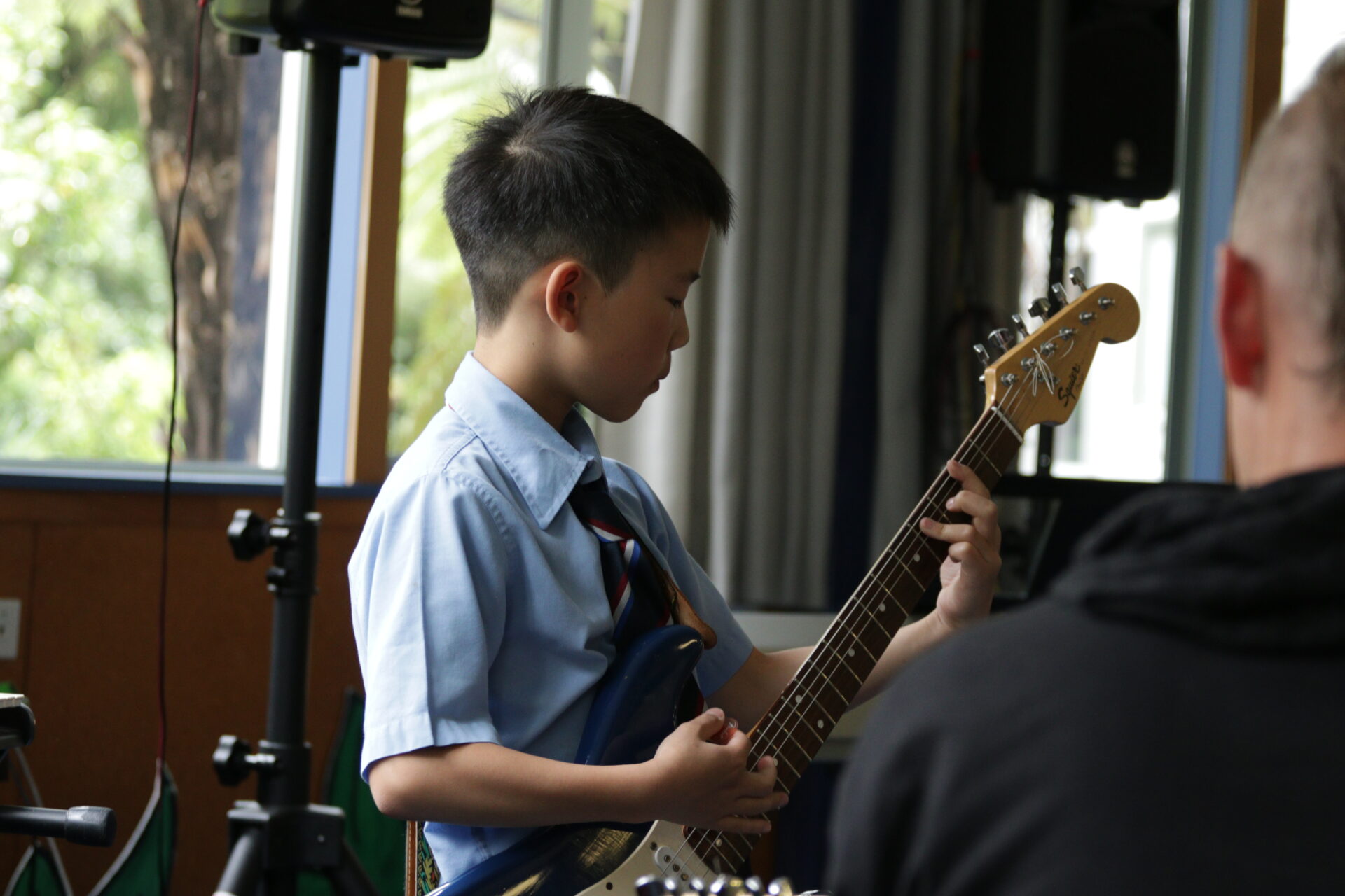 A young boy in a blue school uniform shirt and tie plays an electric guitar indoors, focused on his performance, with blurred people and musical equipment in the background