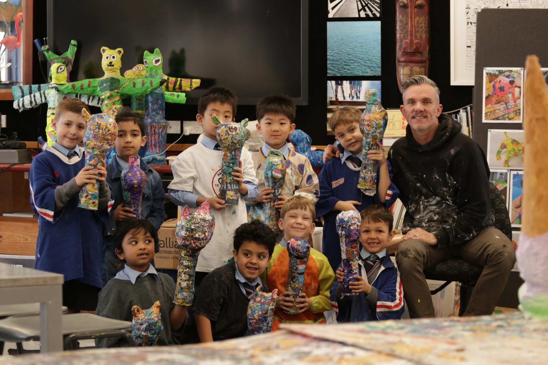 A group of smiling children and their teacher pose in an art classroom, each holding colorful papier-mâché sculptures. Art projects and creative decorations fill the background, creating a lively, creative atmosphere