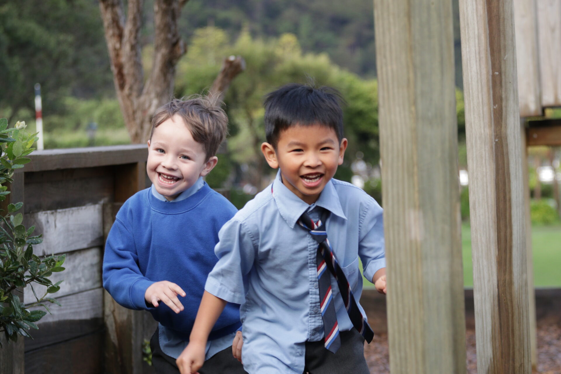 Two boys in school uniform run and smile near the wooden playground at Wellesley College