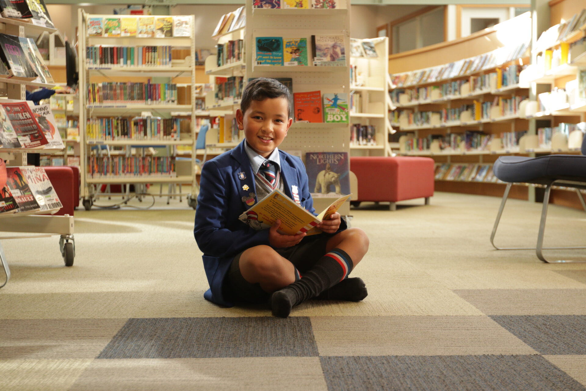 A young boy in a school uniform sits cross-legged on the library floor, smiling while holding an open book. Bookshelves filled with books surround him in the well-lit library