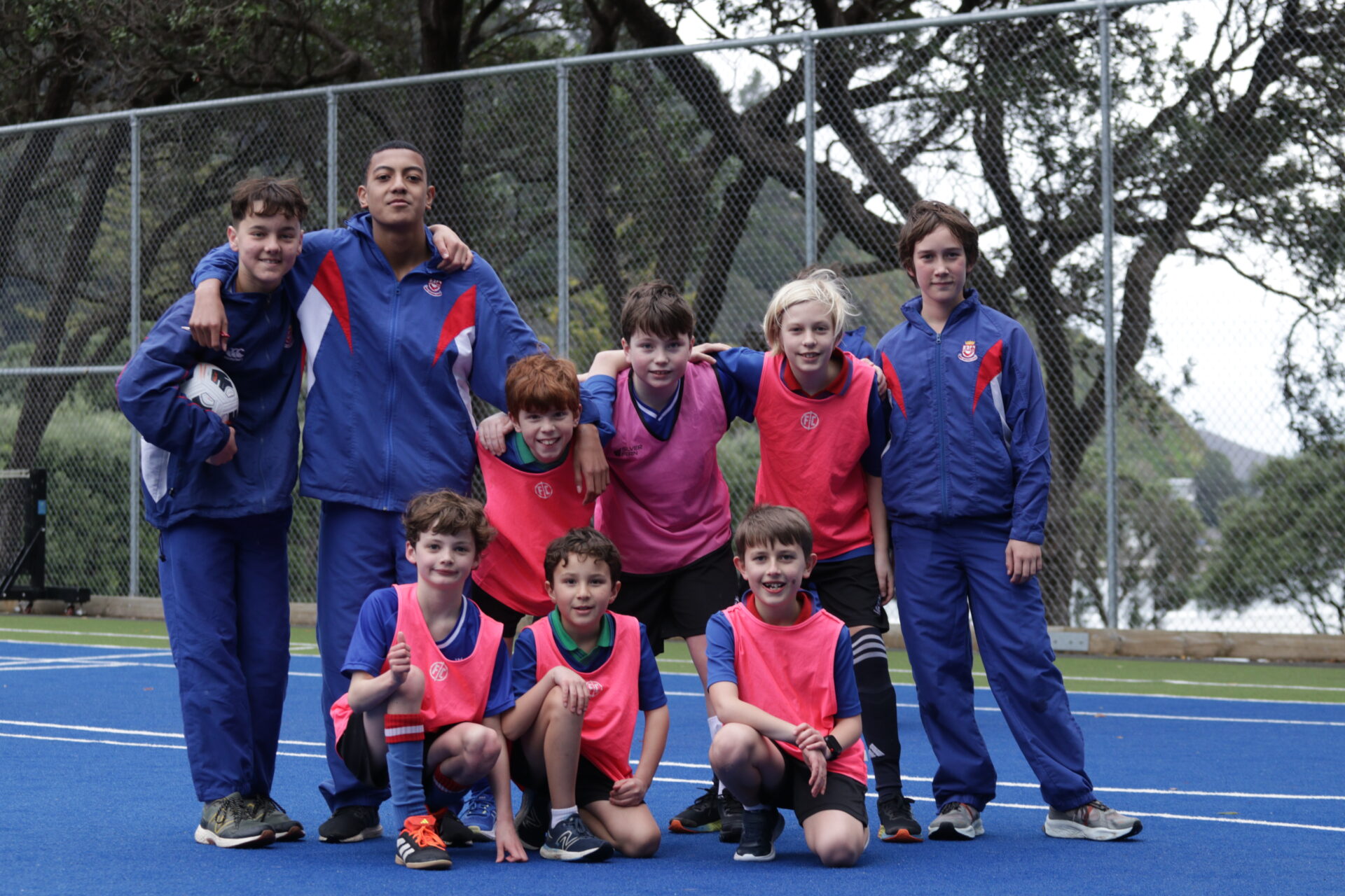 A group of nine boys poses on an outdoor blue sports court. Four boys stand in the back, while five kneel or sit in the front. They wear sports uniforms with pink bibs and look happy, with trees and a fence in the background