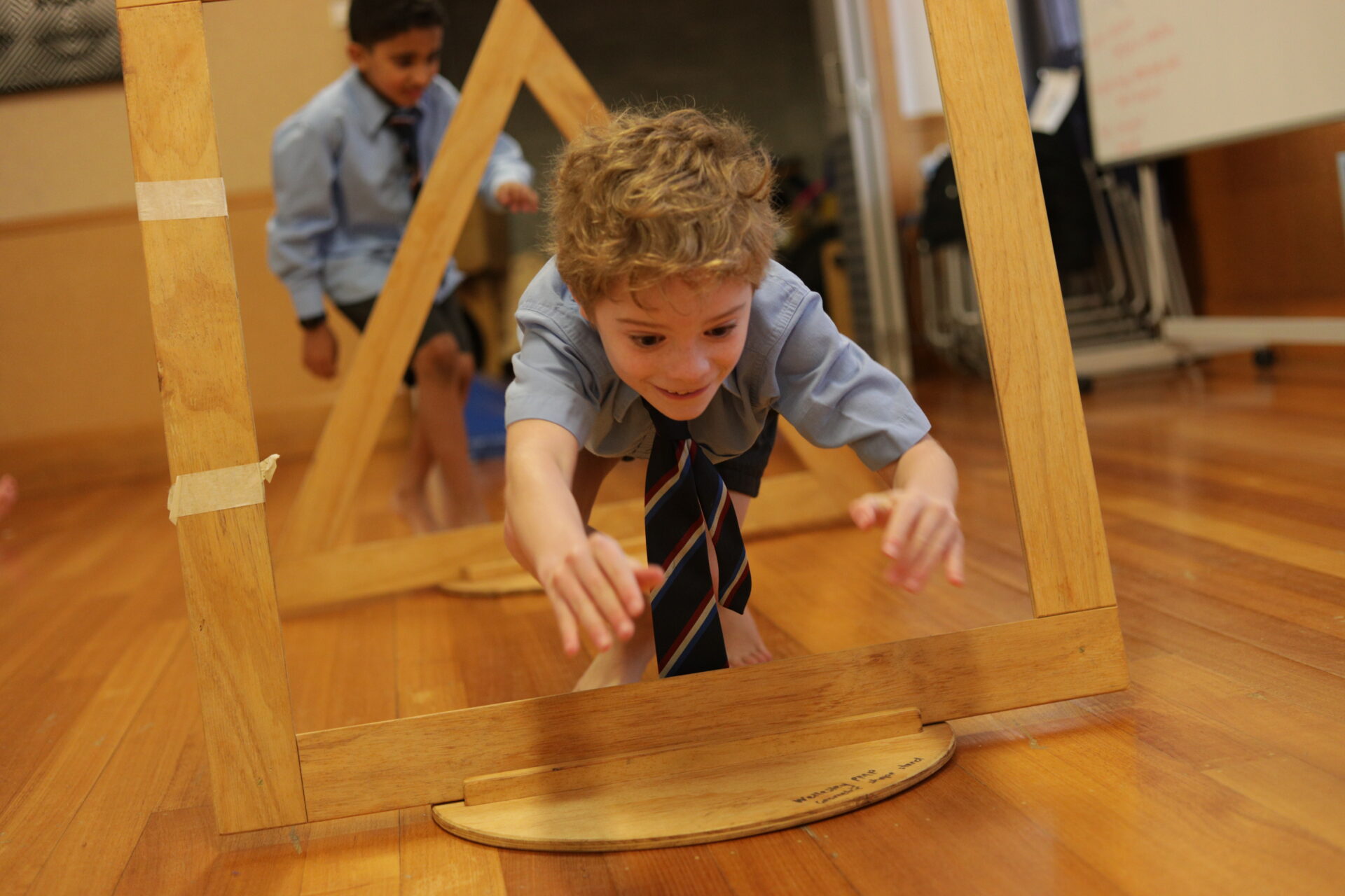 A young boy in a school uniform crawls through a wooden triangular obstacle on a gym floor, smiling, with another boy in uniform following behind him